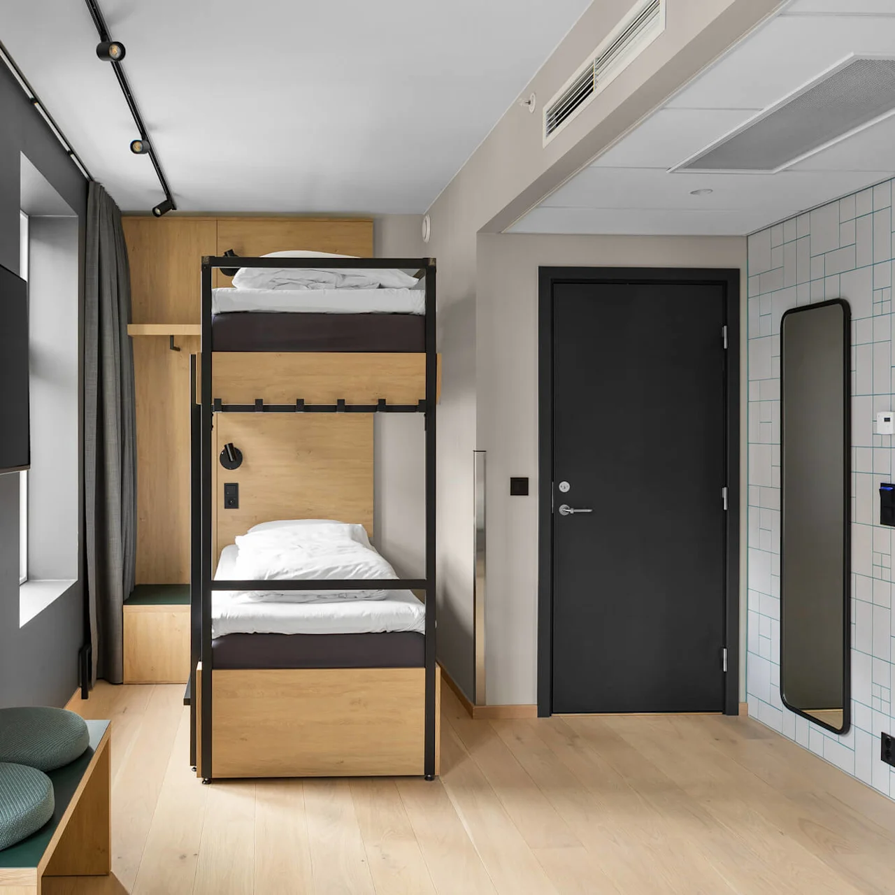 Bunk bed sits in a modern bedroom; room features a wall-mounted TV, two green cushions on a bench, a mirror next to a black door, and a sleek, minimalistic design.