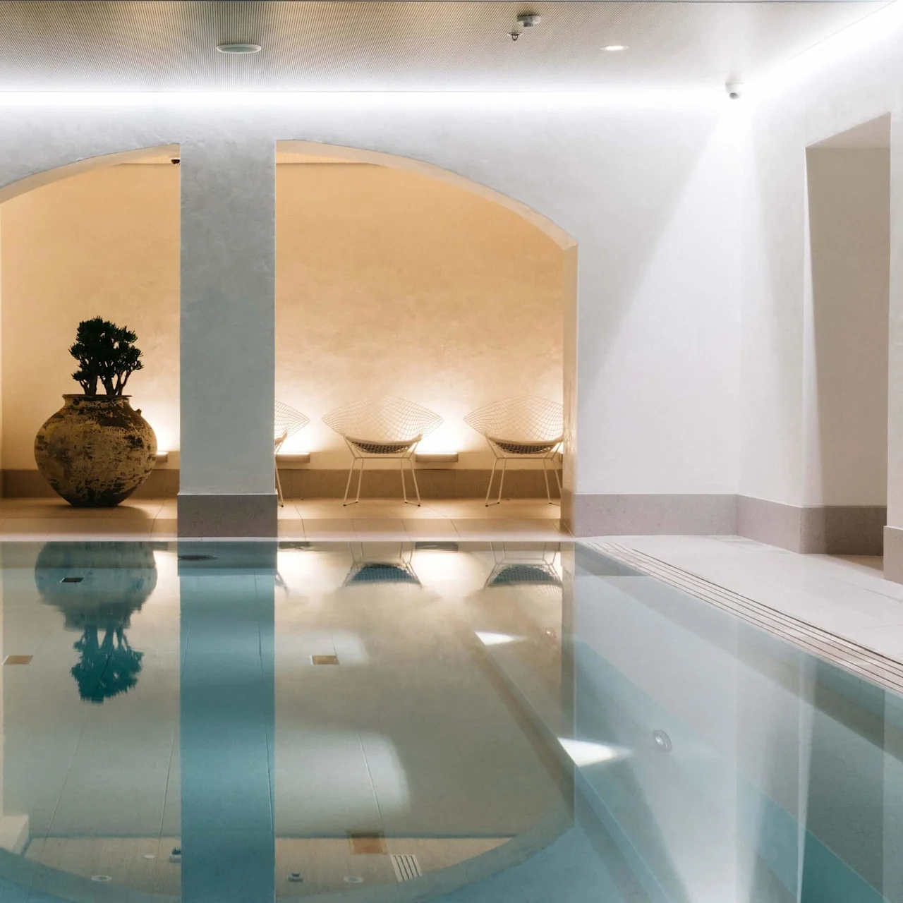 Et indendørs pool reflekterer en alkove med stole og en stor krukkeplante i et roligt, veloplyst rum.