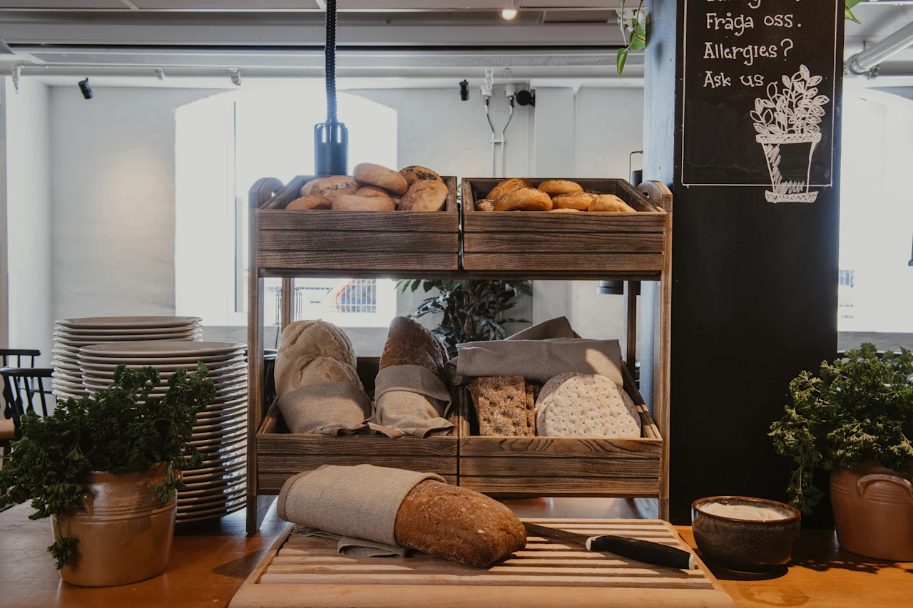 Rustiikkinen leipäesillepano, jossa on erilaisia leipiä, sämpylöitä ja näkkileipää, lautasten ja kyltin kanssa, jossa lukee 'Fraga oss. Allergies? Ask us'.