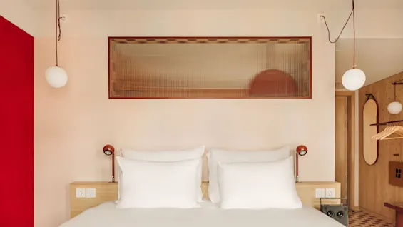 A modern and cozy hotel room with a neatly made bed, white linens, and stylish lighting. A red accent wall adds a pop of color.