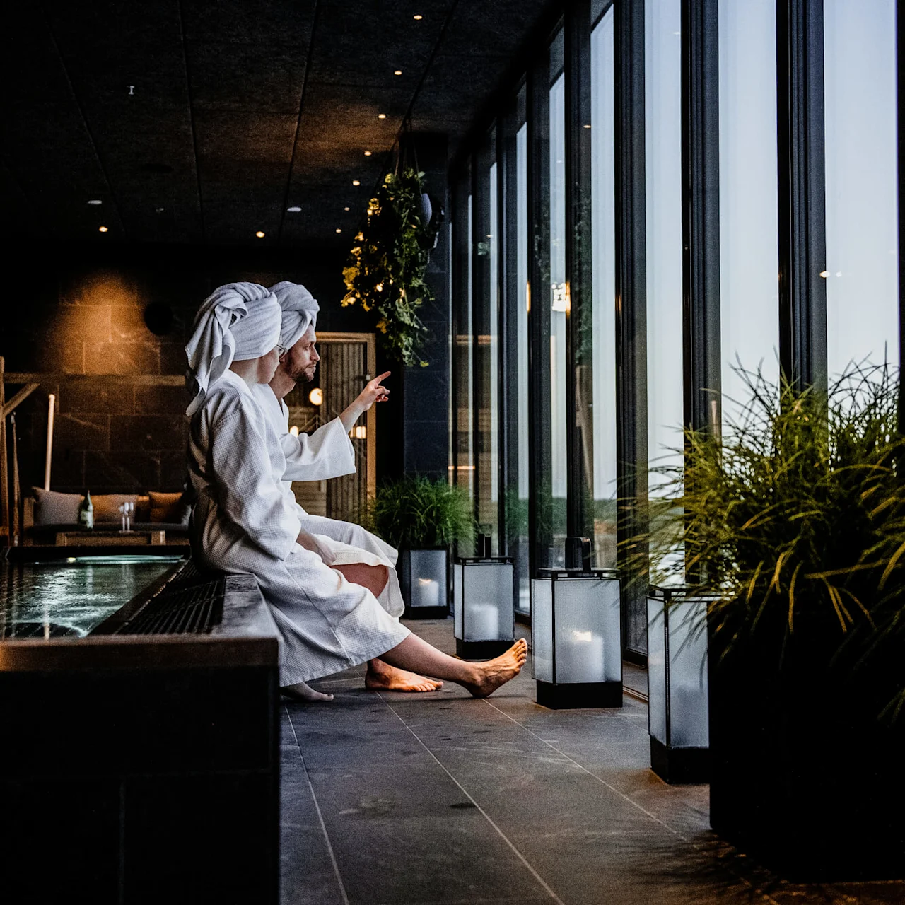 To personer i hvide badekåber sidder ved en pool og kigger ud gennem store vinduer. En person gestikulerer mod ydersiden. Grønne planter og lanterner dekorerer det spa-lignende interiør.