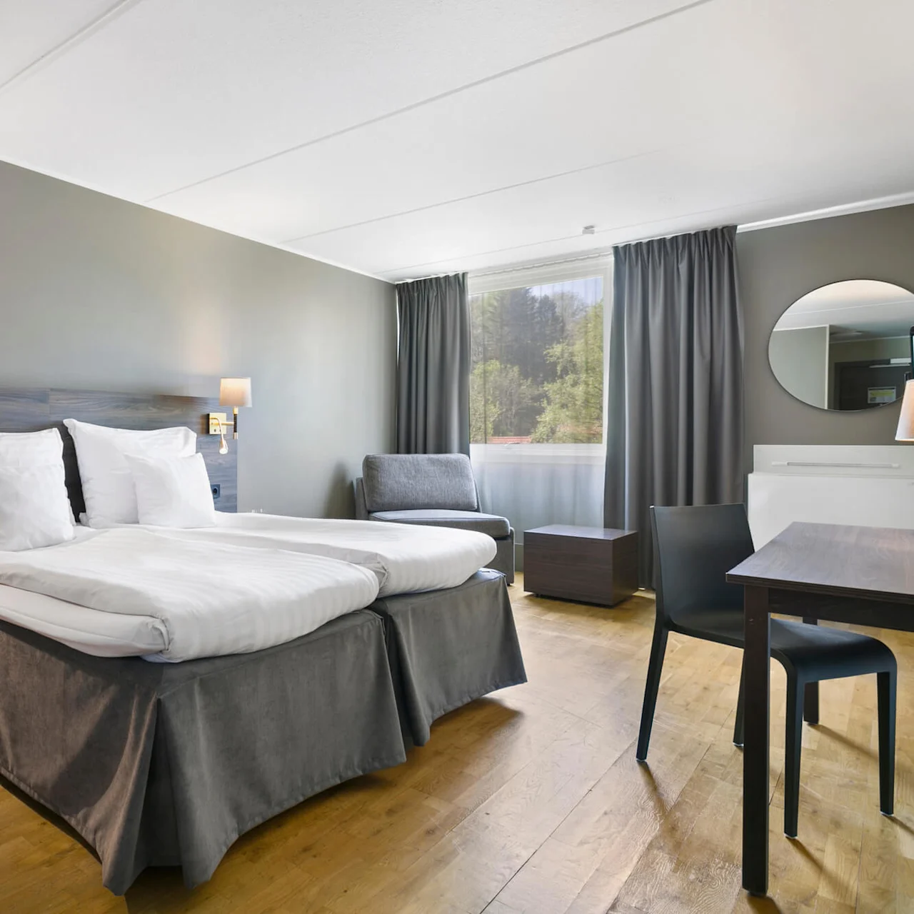 En seng med hvidt sengetøj står ved siden af et lille bord med lampe i et moderne hotelværelse. Stort vindue med gardiner og skovudsigt, stol og skrivebord i baggrunden.
