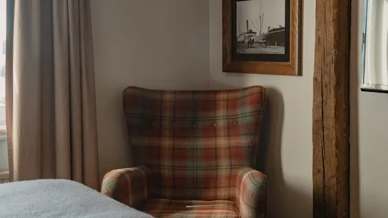 Et varmt og indbydende hjørne af et værelse med en ternet lænestol, en seng og et indrammet nautisk print.