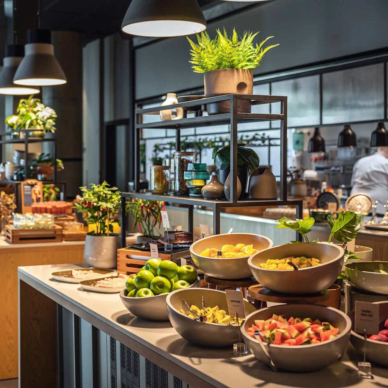 Skåle med frisk frugt vises på en buffet, herunder grønne æbler og vandmelonskiver. Potteplanter og moderne indretning forbedrer det omgivende lys i restaurantmiljøet.