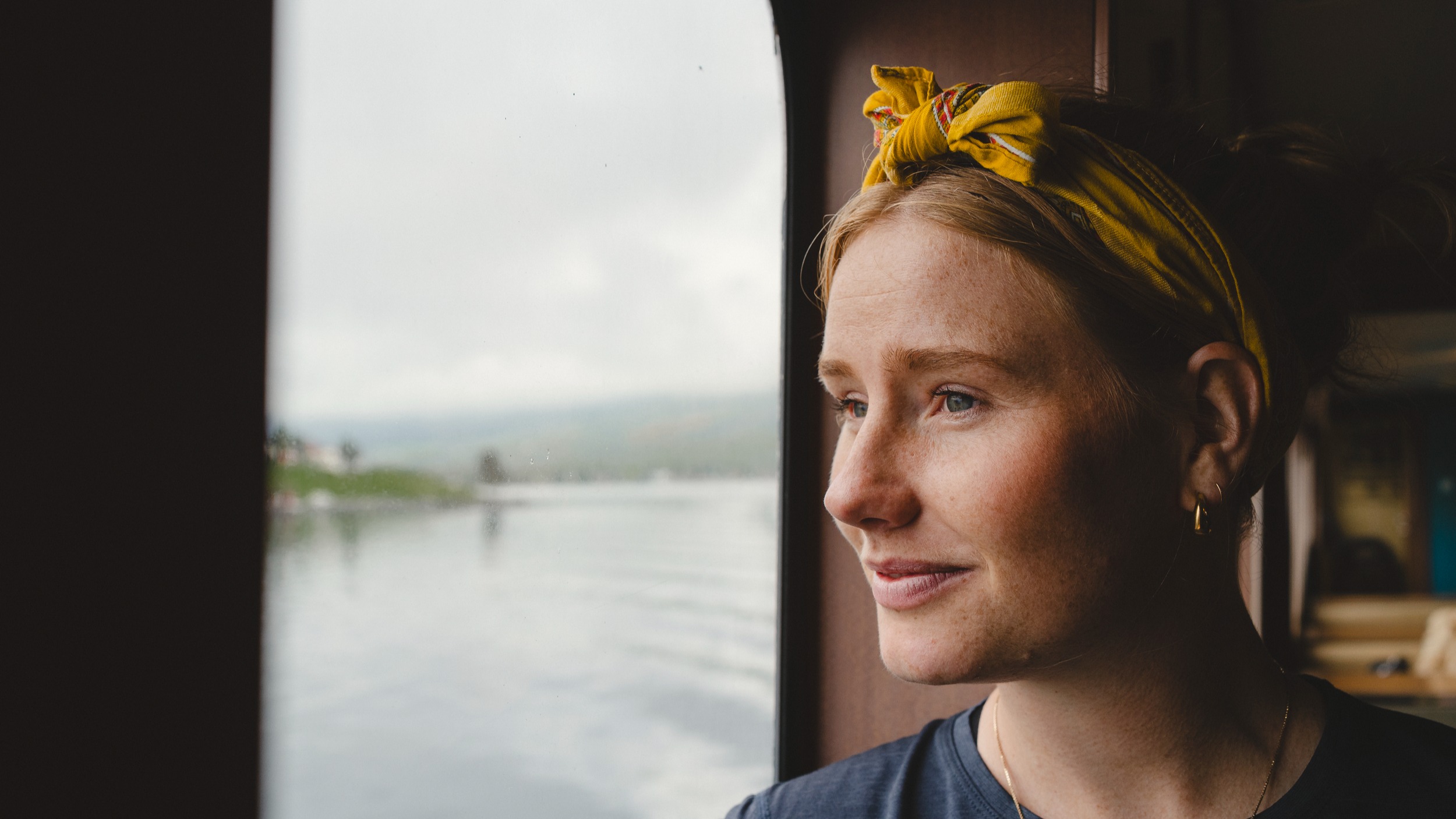 A person with a yellow headband looks out a window at a calm body of water and distant land.