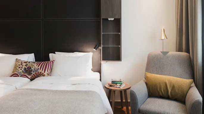 En ryddig seng med hvite puter og en dekorativ pute, ved siden av en lenestol med en gul pute, i et moderne rom med mørke paneler og et lite bord.