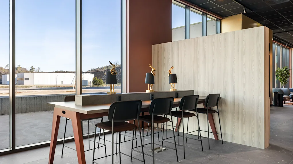 Moderne hotellobby med et langt høyt bord og barkrakker ved store vinduer, som tilbyr en lys arbeidsplass.