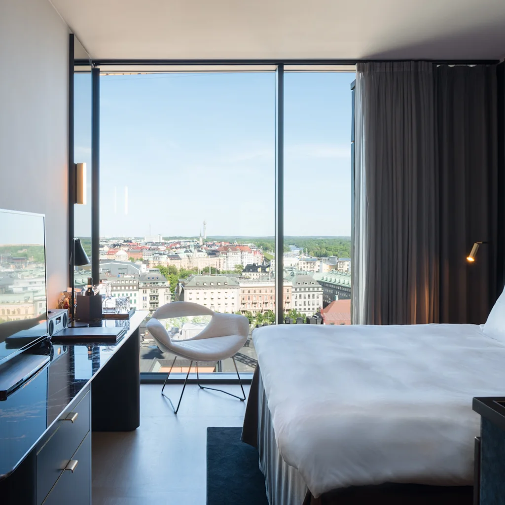 Et lyst hotellrom med en fantastisk byutsikt fra et gulv-til-tak-vindu, komplett med en koselig seng og et elegant skrivebordsområde.