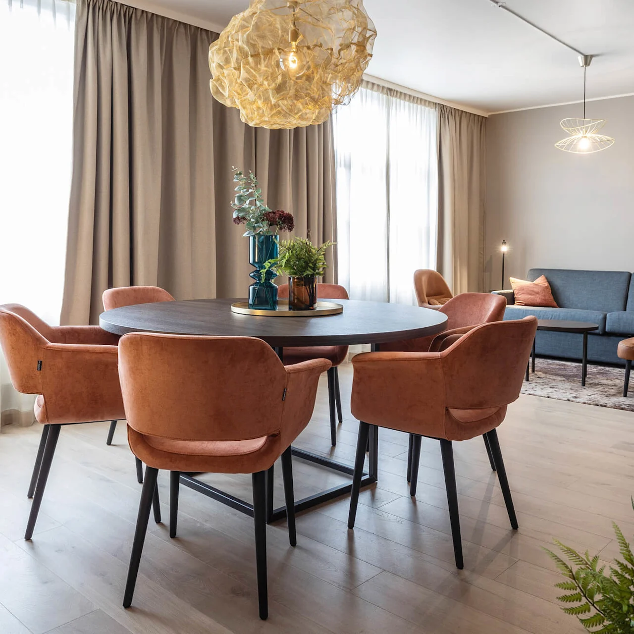 Spisebord omgivet af orange stole, pyntet med vaser, placeret i en moderne stue. Sofa og lænestole komplementerer rummet, oplyst af dekorative pendellamper.