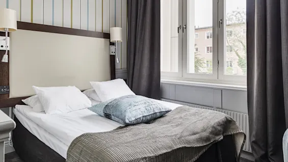 A neatly made single bed against a striped wall, with two lamps, near a large window with sheer and dark curtains, in a well-lit room.