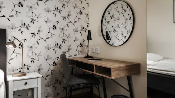 Ett skrivbord i trä med en svart lampa och en anteckningsbok står intill en blommig tapetserad vägg, under en rund spegel. I närheten står ett nattduksbord med en lampa.