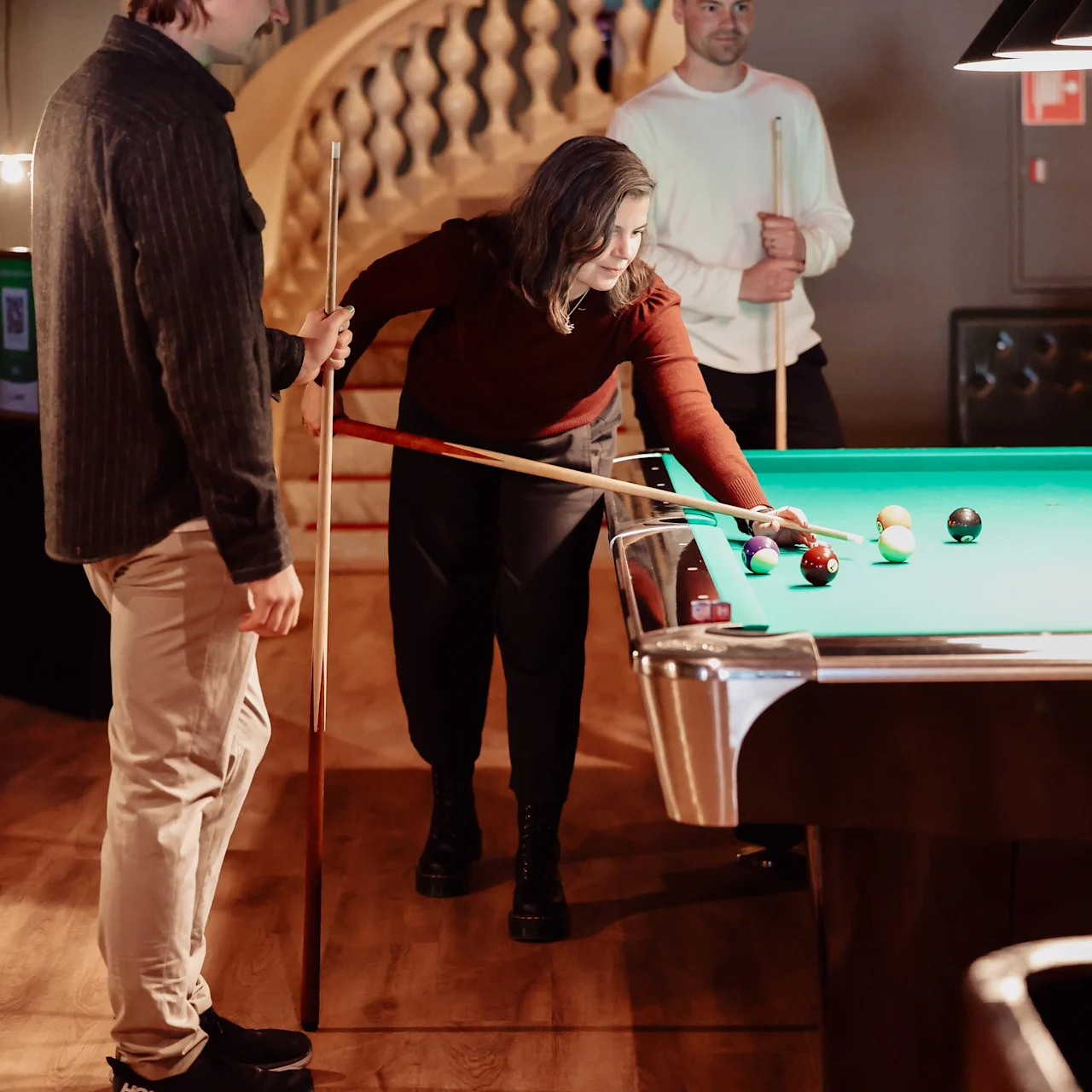 A woman leans over a pool table, aiming a cue at the balls, while two men watch in a warmly lit room with a curved staircase in the background.