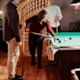 A woman leans over a pool table, aiming a cue at the balls, while two men watch in a warmly lit room with a curved staircase in the background.