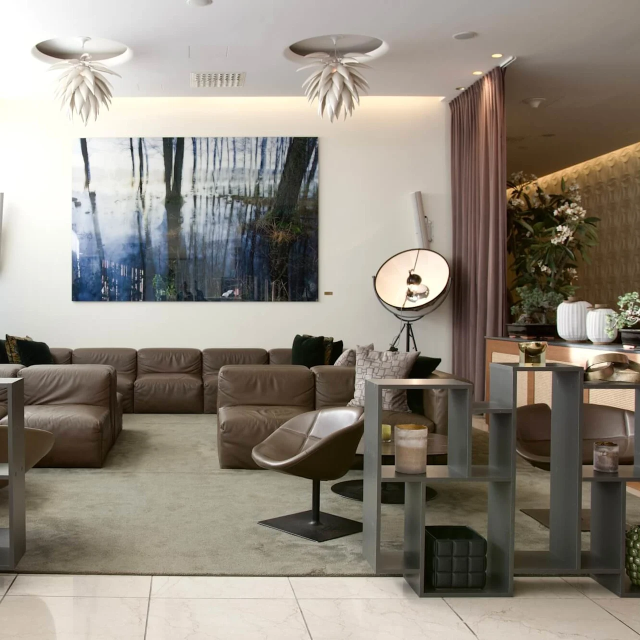 A brown leather sofa sits in a modern lounge adorned with abstract wall art and decorative plants, illuminated by hanging lights and natural daylight through a large window.