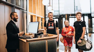 People smiling in a modern hotel lobby, one at the reception desk and three others entering, dressed in sportswear.