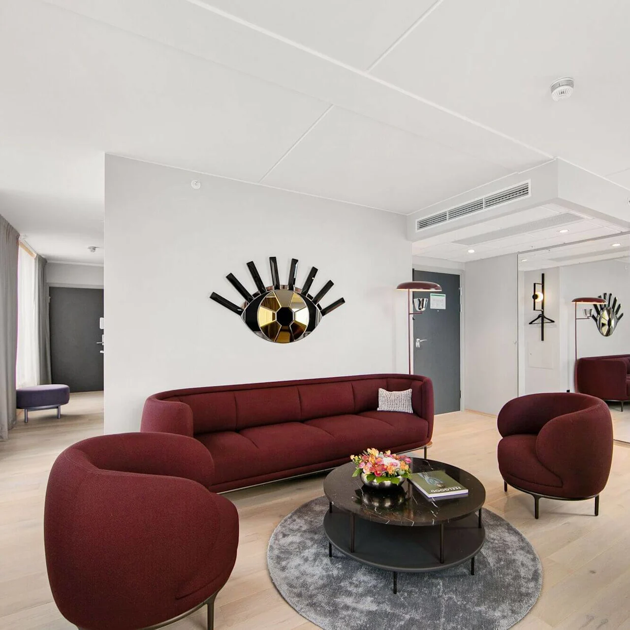 A burgundy seating arrangement with two chairs and a sofa surrounds a round coffee table and rug in a modern, spacious living room with large windows and a wall mirror decoration.