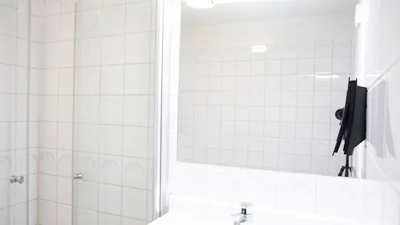 Et badeværelse med hvide fliser har en glassdør til bruseniche og et stort spejl over en vask med vandhane. En sort genstand hænger på væggen.