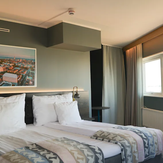 A neatly made bed with white pillows and patterned blankets sits in a hotel room. A cityscape photo hangs above, with large windows and curtains to the right.