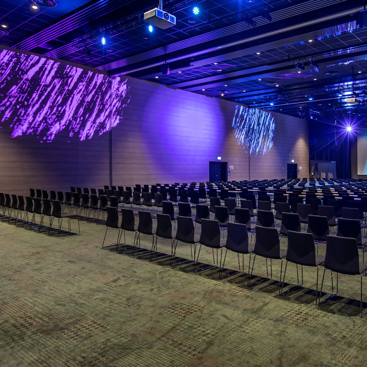 A large conference hall features rows of empty chairs facing a stage. Colorful lights in purple, blue, and pink illuminate the stage and side walls, creating a vibrant atmosphere.