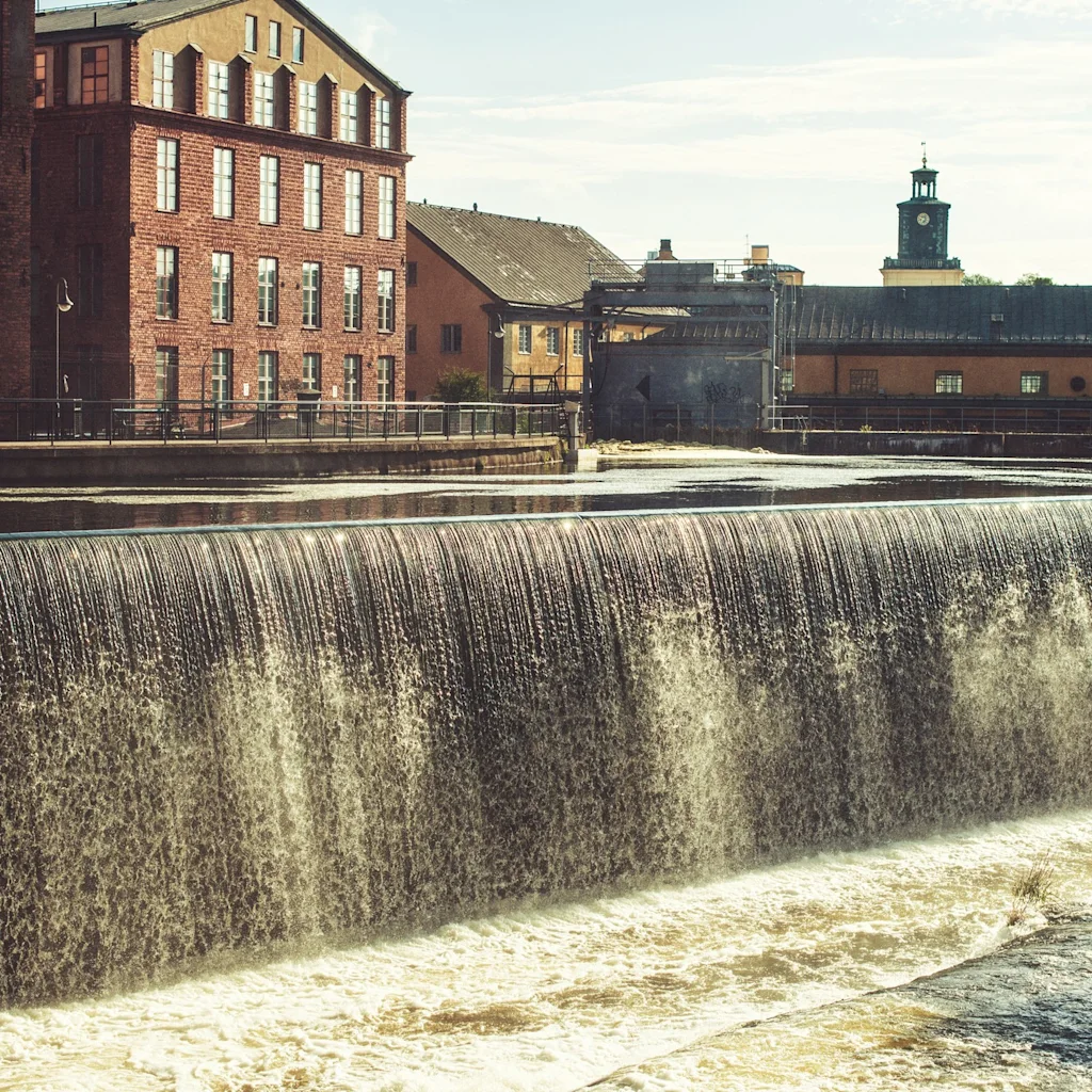 A wide waterfall or dam with old brick industrial buildings and a clock tower in the background. Industrilandskapet in Nörrköping, Sweden.