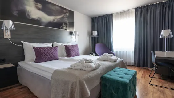 Ett prydligt arrangerat hotellrum med en dubbelsäng med två lila kuddar och vita morgonrockar. En turkos ottoman står vid fotändan, med ett skrivbord och stol i närheten.