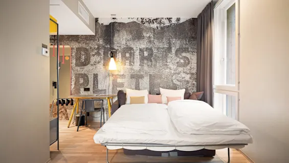 A fold-out bed with white bedding extends in a cozy room, featuring a rustic wall with the text "DROP ART'S PLEATS." A wooden desk and a hanging lamp add warmth.