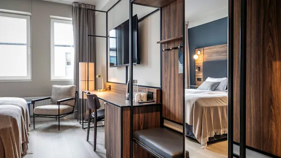 Ett modernt hotellrum med en bekväm säng, ett skrivbord av trä med lampa och koppar, en TV på väggen och en stoppad stol, vilket skapar en mysig atmosfär.