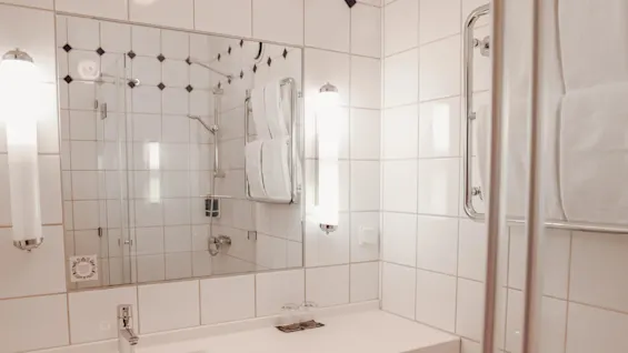 Sink with chrome faucet sits below a large mirror in a tiled bathroom. Towels hang on racks, and two glasses rest on the counter.