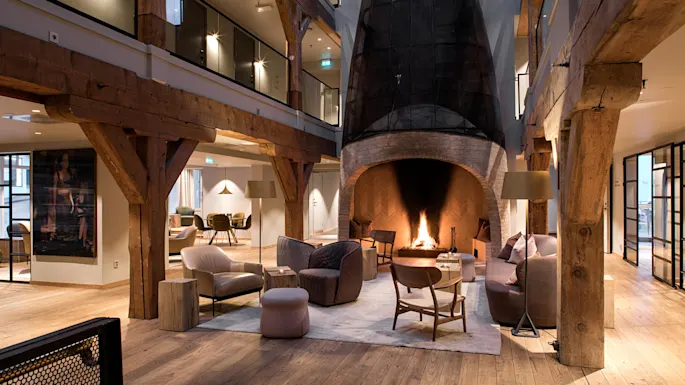 A fireplace crackles warmly, surrounded by modern seating in a rustic lounge. Timber beams accent the spacious two-story interior, filled with soft lighting and inviting decor.