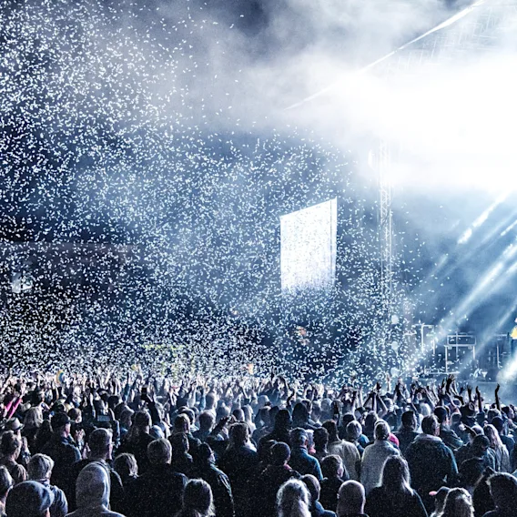 En stor folkemengde på en konsert med sterke scenelys og konfetti som faller, noe som skaper en energisk atmosfære.