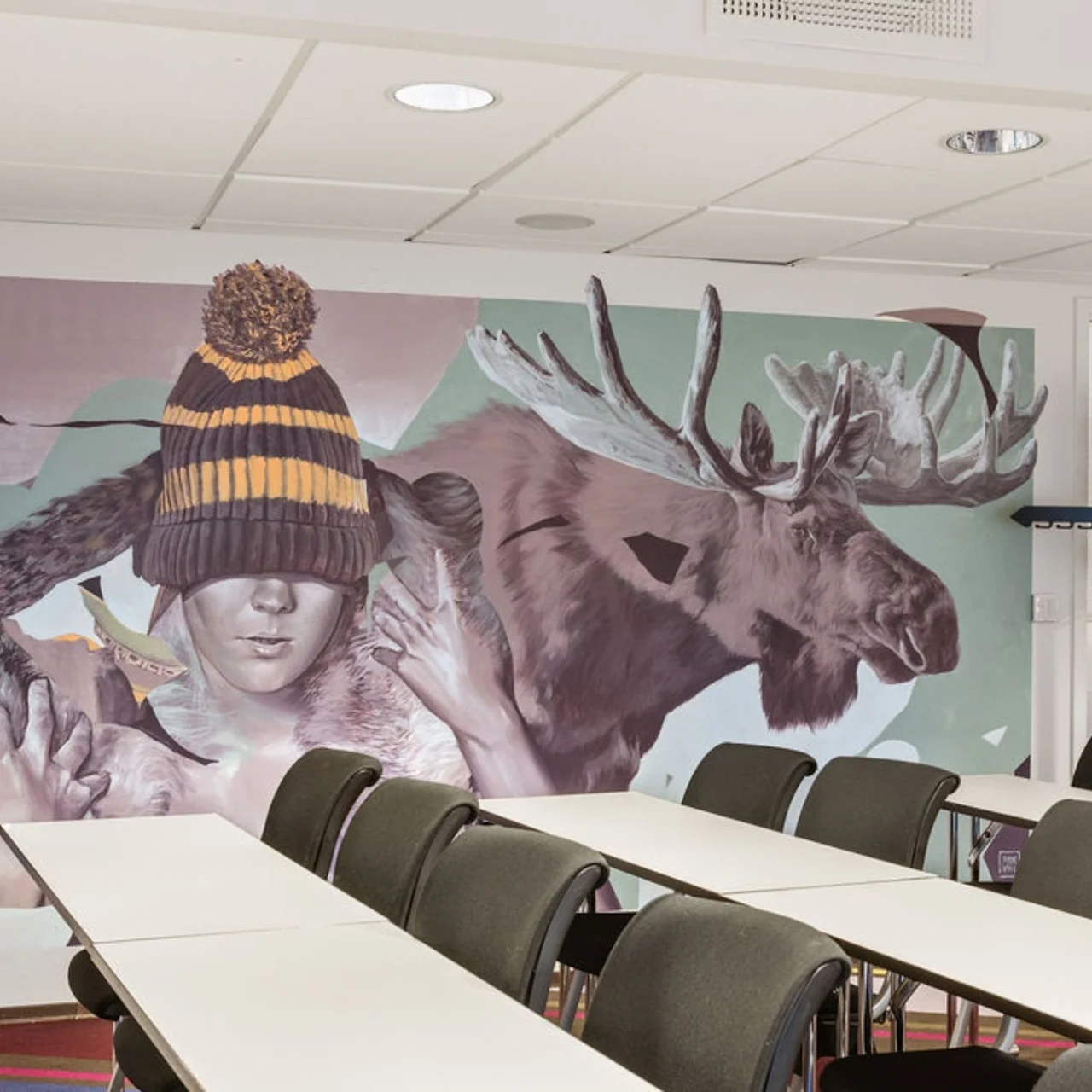 A mural of a child with a beanie and a moose is painted on the wall. Classroom desks with chairs are arranged in front of the colorful artwork.