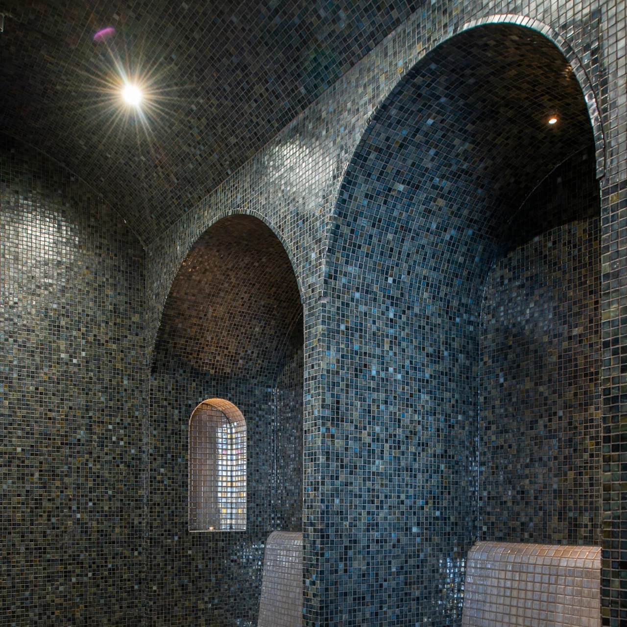 Mosaic-tiled benches rest under archways in a dimly lit steam room with a tiled floor and small window, creating a serene and intimate atmosphere.