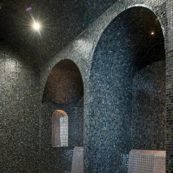 Flerfärgade mosaikklädda bänkar under valv i ett svagt upplyst ångrum med kakelgolv och ett litet fönster, vilket skapar en rofylld och intim atmosfär.