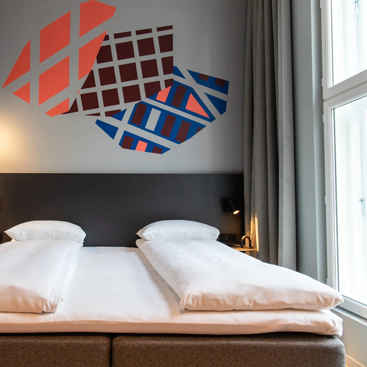 Bed with white linens, flanked by modern lamps, stands against a wall featuring abstract geometric artwork; large window with curtains on the right.