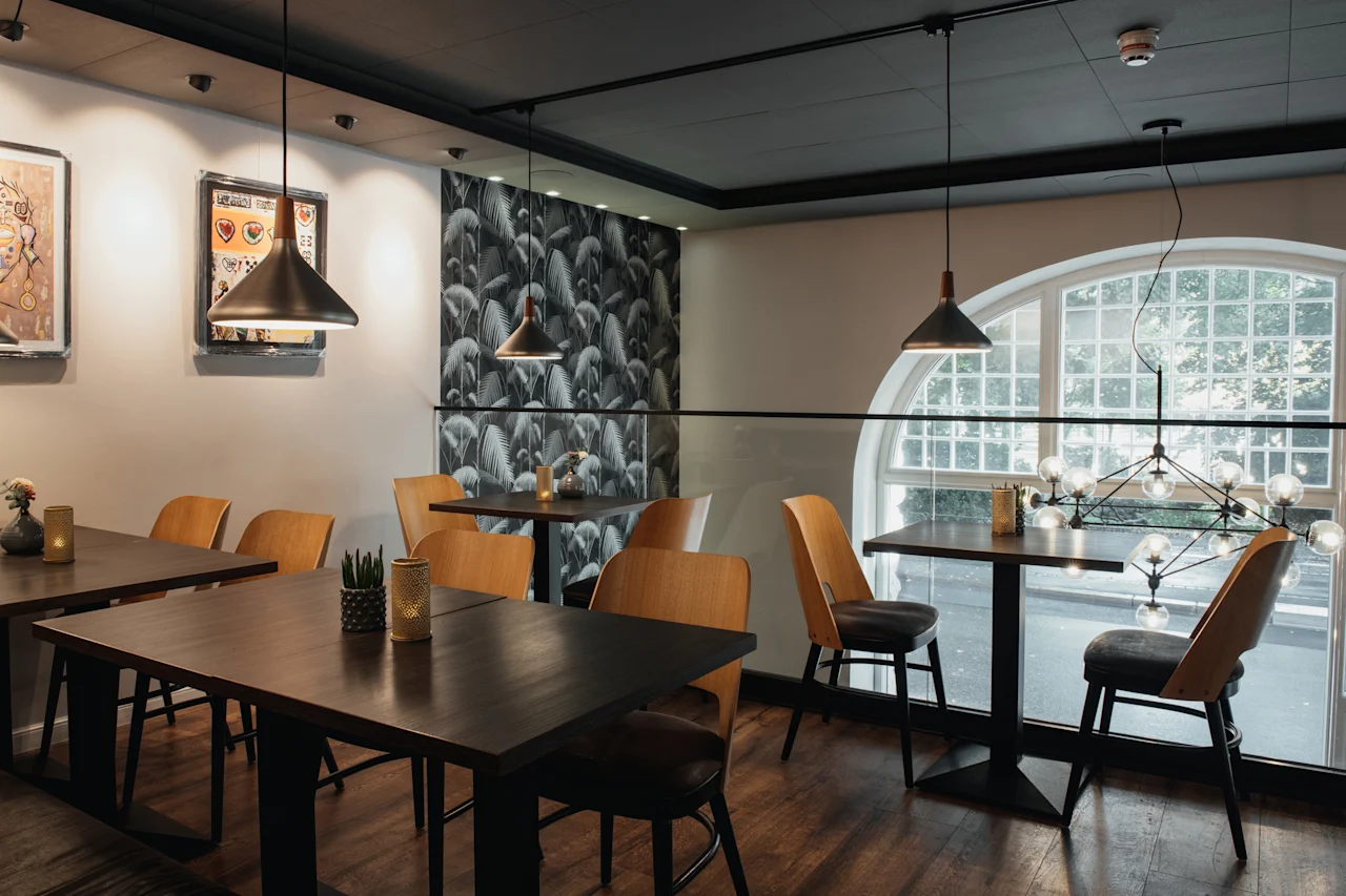 Et moderne restaurantinteriør med mørke træborde, stilfulde stole og unikke belysningsarmaturer, med et stort buet vindue.