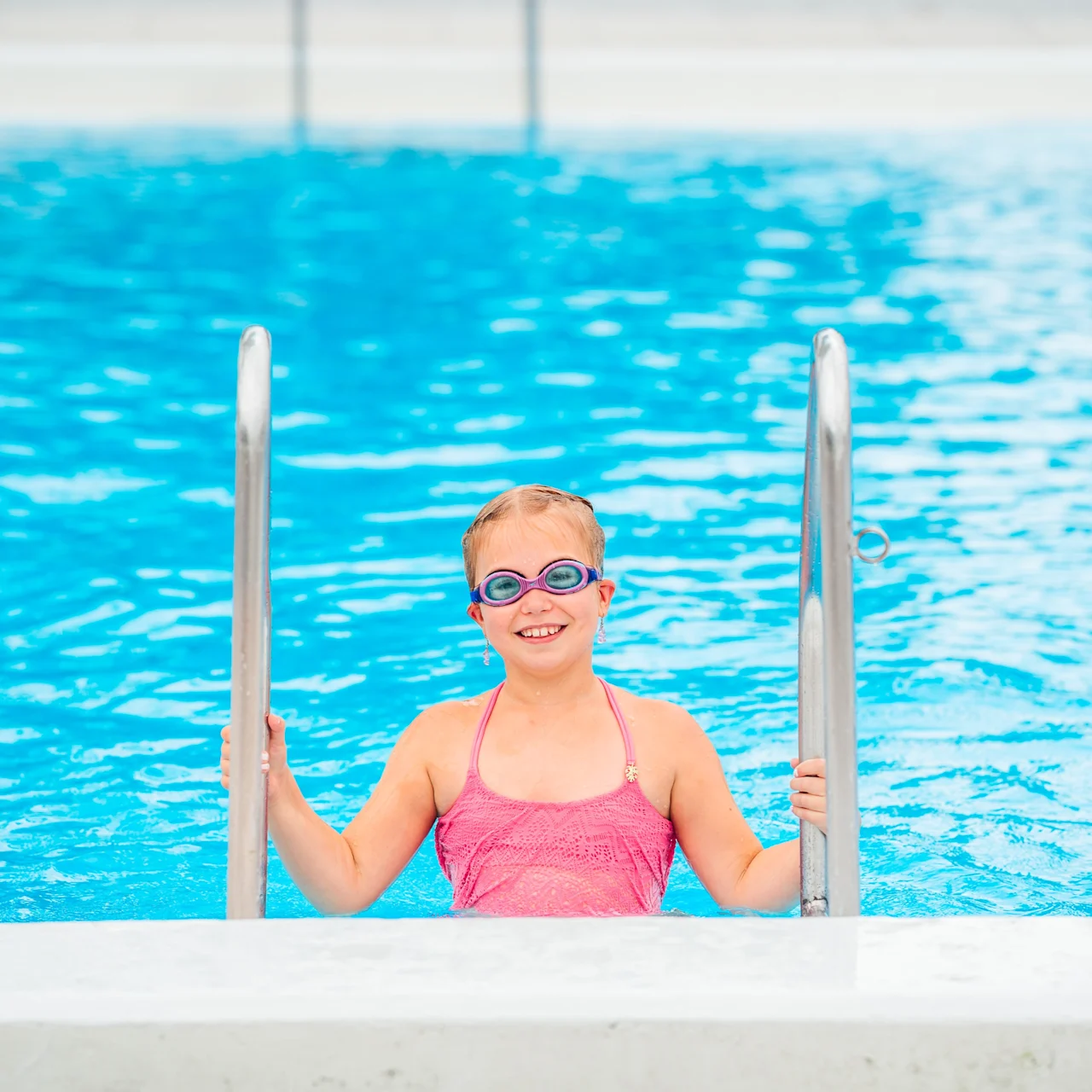 Child wearing goggles smiles while holding pool ladder; standing in a bright blue swimming pool.