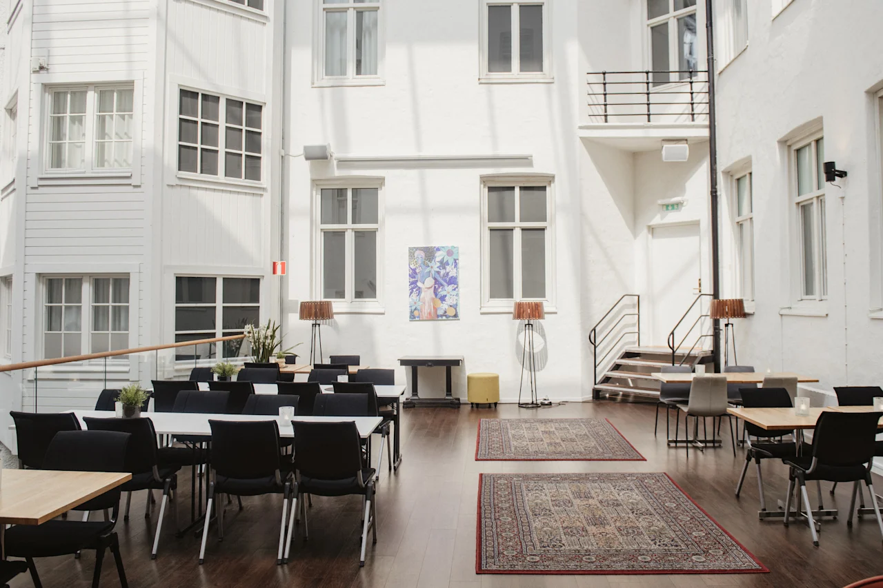 En solbelyst atrium har rektangulära bord med svarta stolar, omgivna av vita väggar med flera fönster. Mattor täcker trägolvet, och konstverk hänger längs bakväggen.