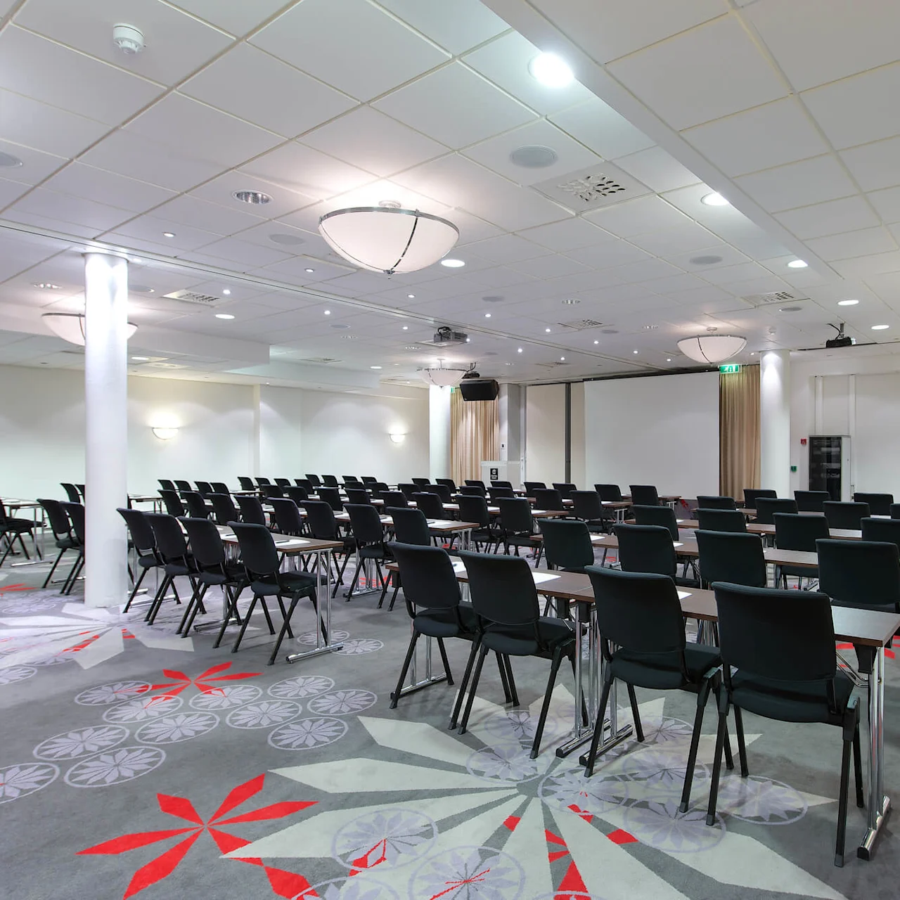Rows of black chairs facing a projector screen in a modern conference room with patterned carpet and ceiling lights.