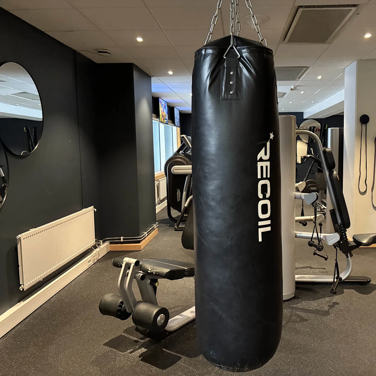 En punching bag merket "RECOIL" henger i et treningssenter, omgitt av treningsutstyr, speil og motstandsbånd.