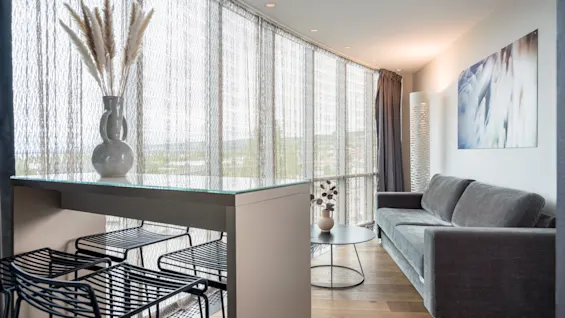 A glass-top table with a vase of dried flowers stands indoors, next to black metal stools, facing a gray sofa by large windows with sheer curtains.