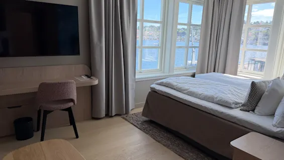 A bright hotel room with a comfortable bed, a desk area, and large windows offering a scenic view of the water.