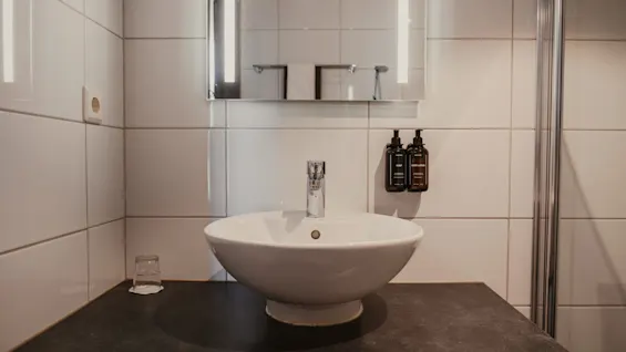 A modern bathroom with a white sink, illuminated mirror, and dispensers for soap and body lotion. A glass sits on the dark counter.