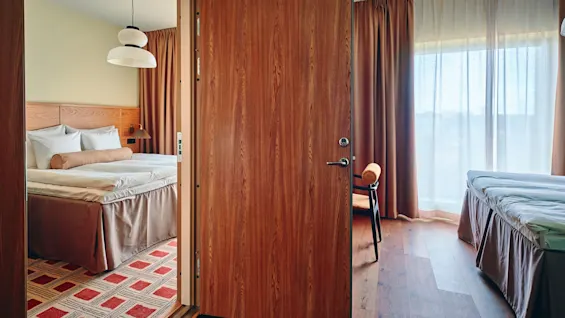 Et koselig hotellrom med seng, mønstret teppe og åpen dør til et annet område med seng og lyst vindu.