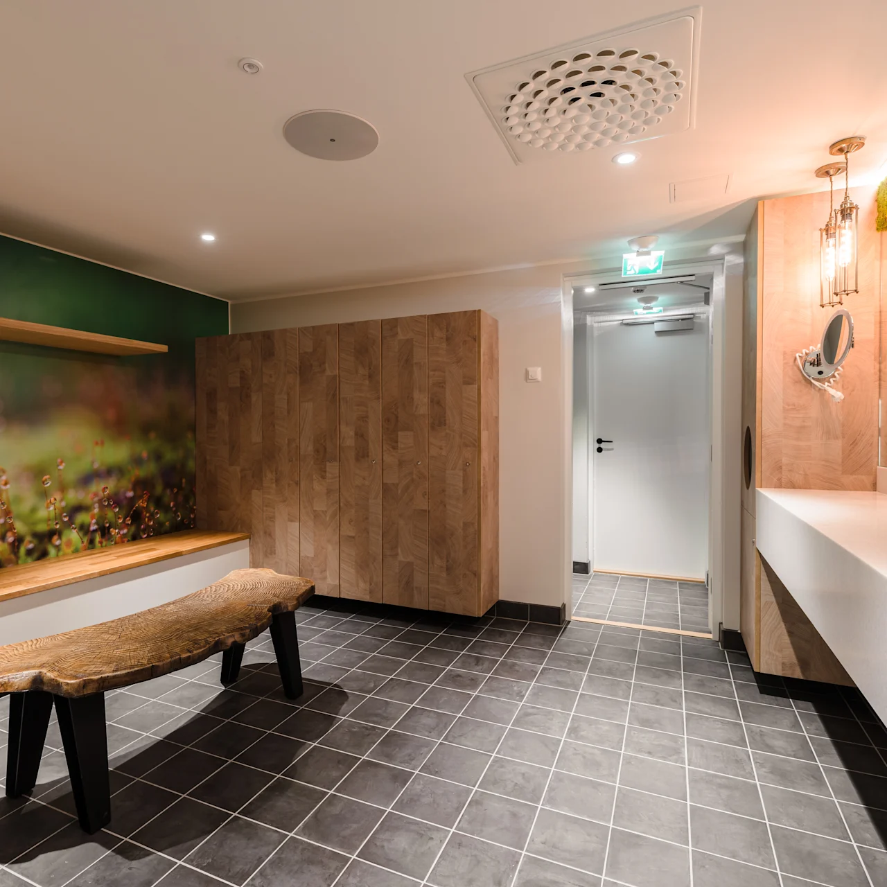 Wooden bench stands on gray-tiled floor, surrounded by nature-themed wall art and wooden panels; bright lights and mirrors reflect modern aesthetic with moss decor overhead. Door leads to an adjoining room.