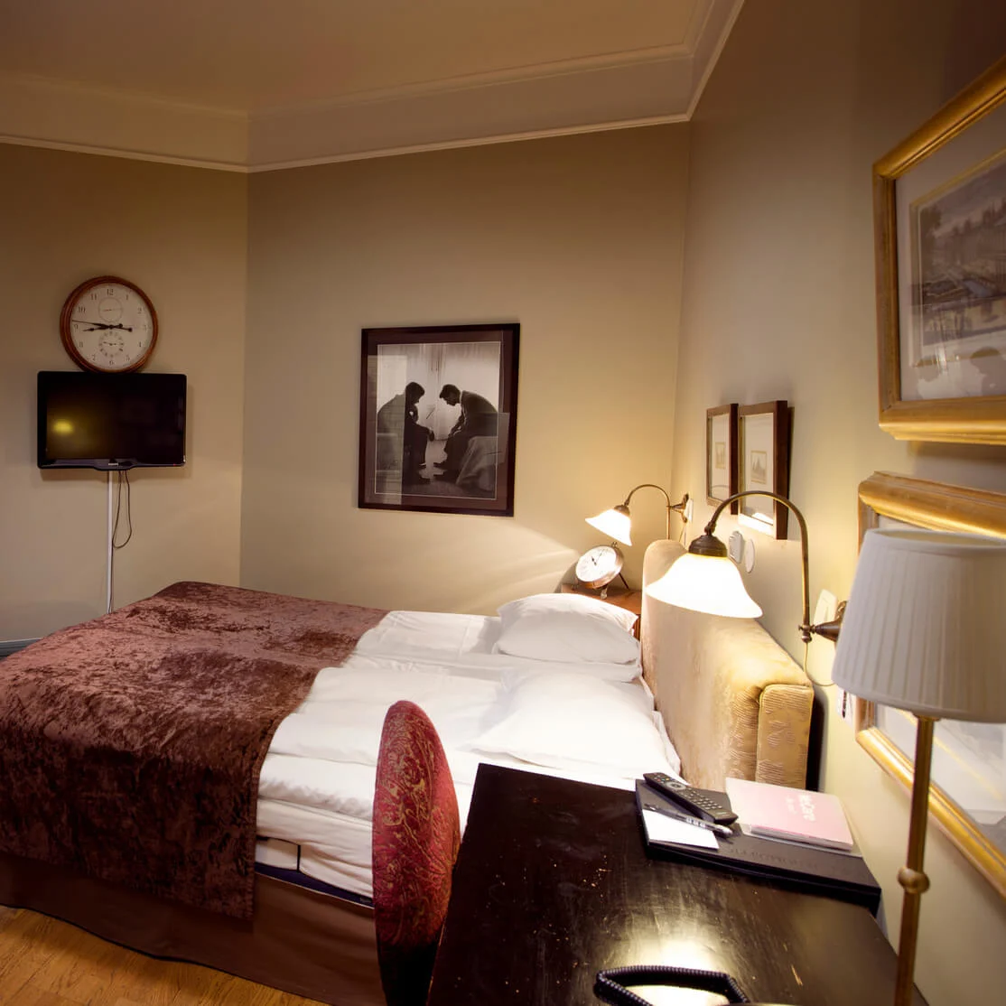 Et godt opplyst hotellrom med en elegant oppredd seng, et sitteområde, en veggmontert TV og innrammede kunstverk, utstråler en koselig og sofistikert atmosfære.