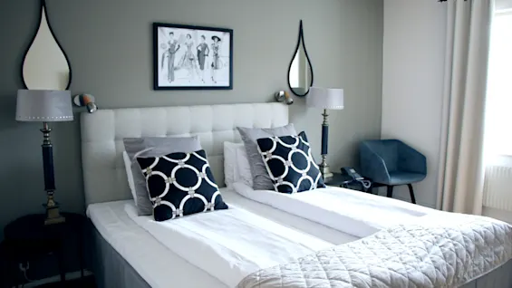 An elegant hotel room featuring a made double bed, patterned pillows, bedside lamps, and stylish mirrors on the wall.