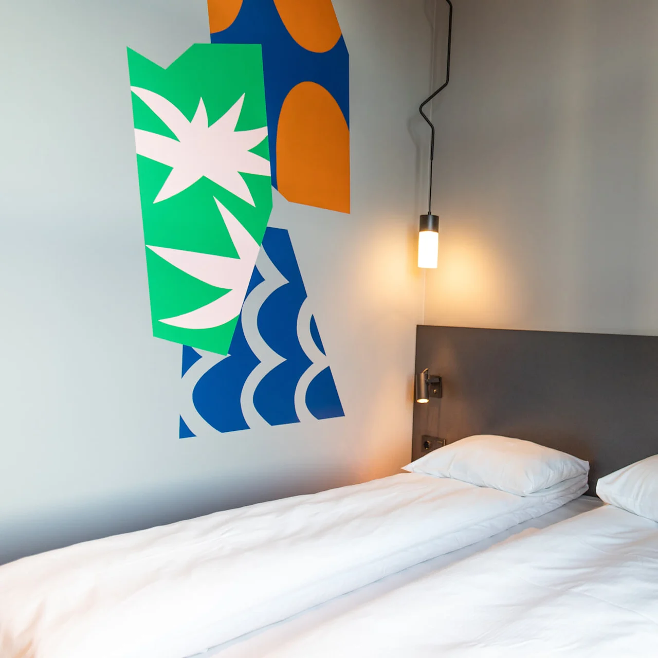 Two neatly-made twin beds rest side by side against a wall; abstract art decorates the wall above, and small lamps provide light, creating a cozy atmosphere.