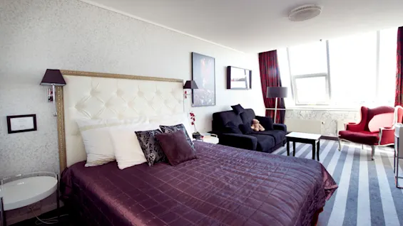 A well-decorated hotel room showcases a large bed with elegant bedding, a sitting area with stylish furniture, and a bright window.