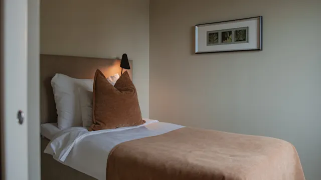 En säng med bruna kuddar och filt står i ett mjukt belyst rum, bredvid en inramad bild på en beige vägg.