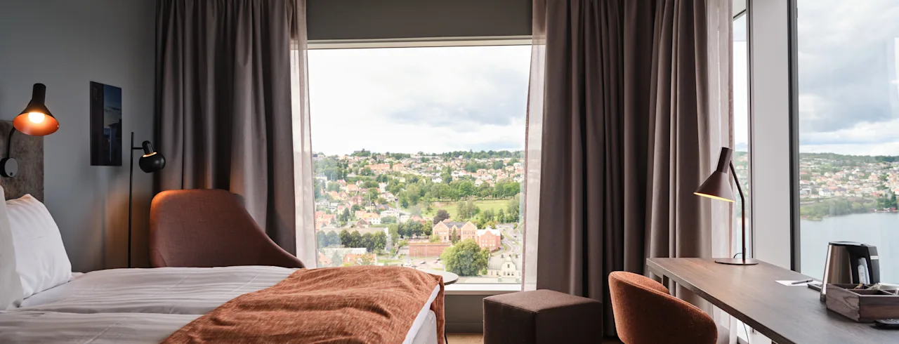 Siististi järjestetty hotellihuone, jossa on sänky, työpöytä ja tuoli. Suuret ikkunat kehystävät näkymää kaupunkimaisemaan, jossa on puita ja rakennuksia.